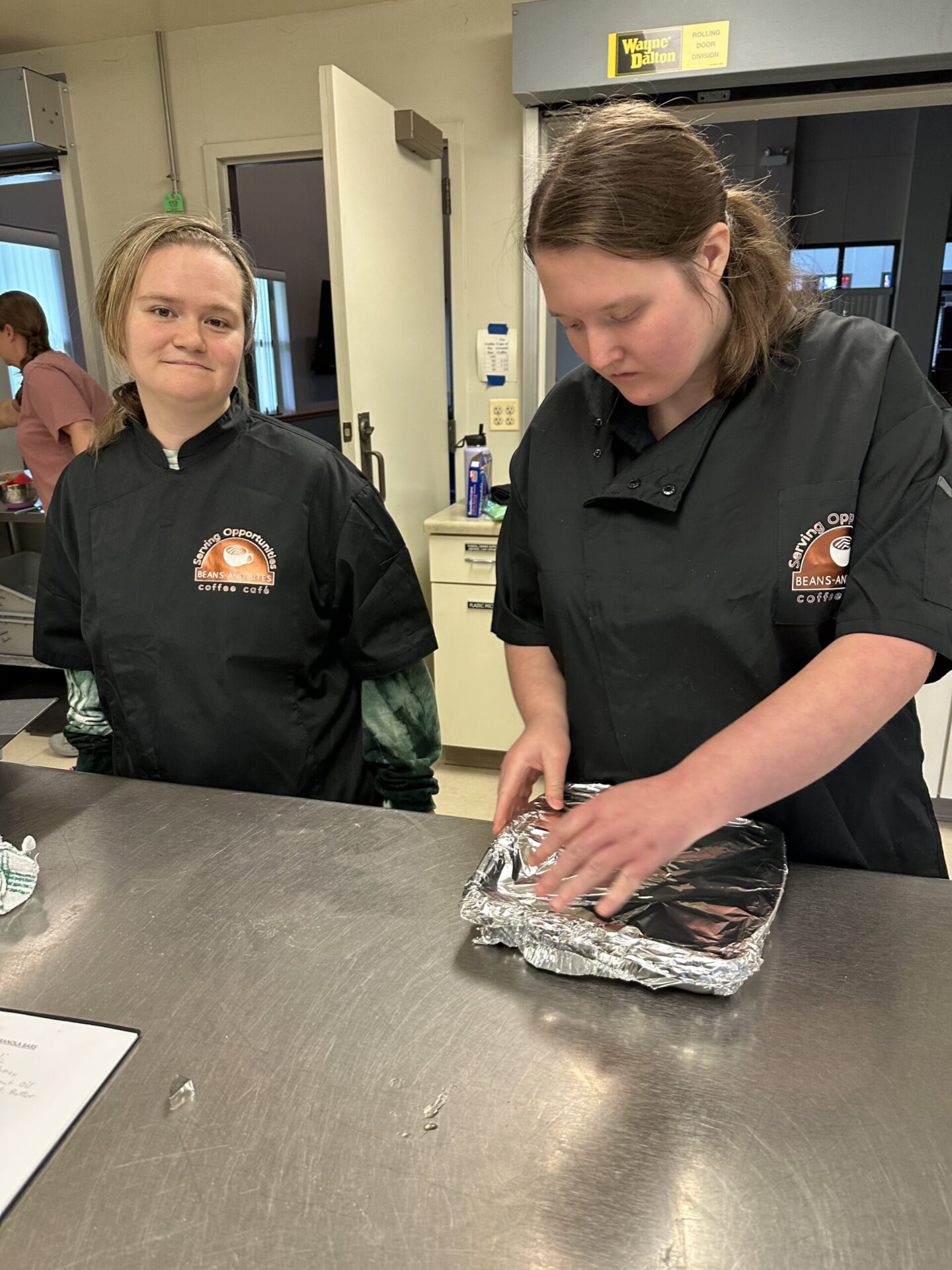Two people in black chef jackets stand at a stainless steel counter in a kitchen. One person is smiling at the camera, while the other is focused on wrapping a tray with aluminum foil.